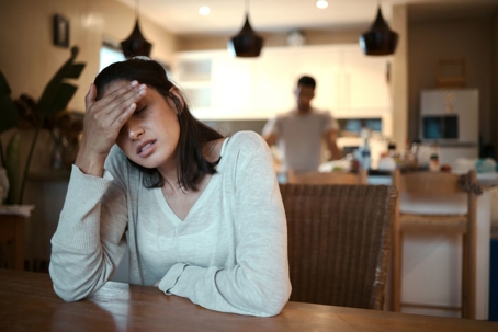 A stressed woman sits at kitchen table while her partner stands in the background