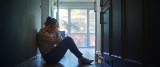 A dark profile shot of abused woman sitting on apartment floor with her head in her hands.