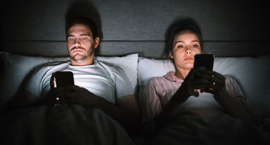 A couple in bed together texts in the dark with screen illuminating their faces.